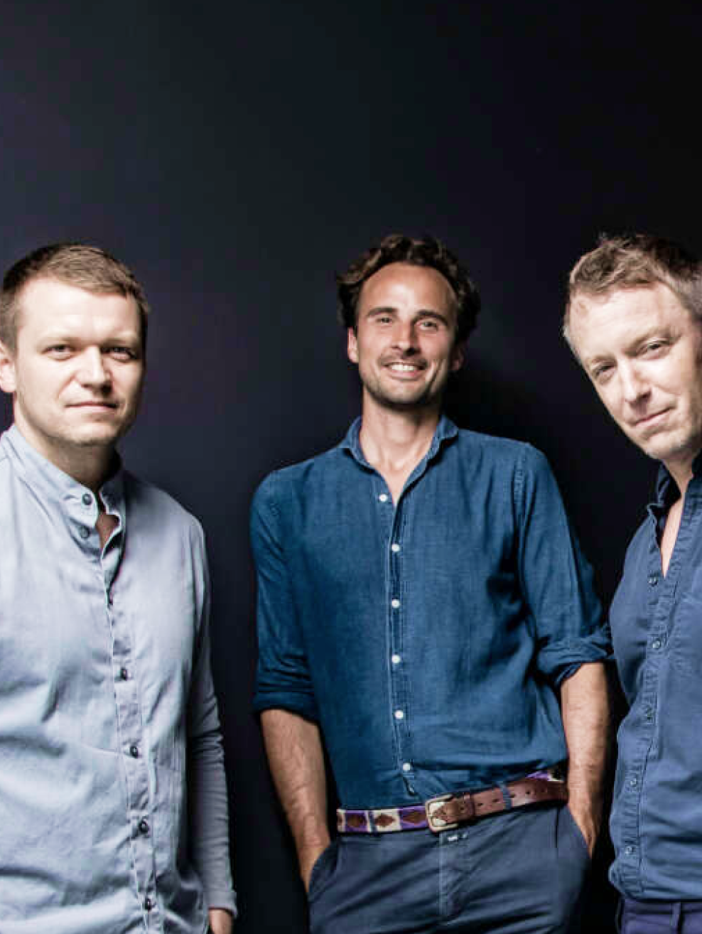 Drei Männer in lässiger Kleidung stehen vor einem schwarzen Hintergrund und blicken in die Kamera.