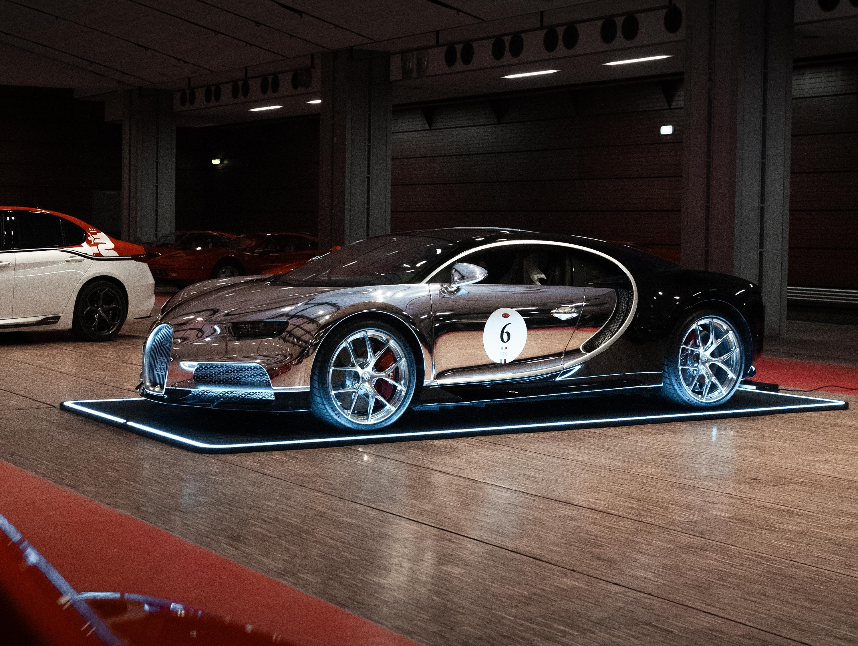 Schwarzer Sportwagen auf beleuchtetem Pad in einer Halle, daneben ein weiteres Auto in Weiß mit rotem Dach.