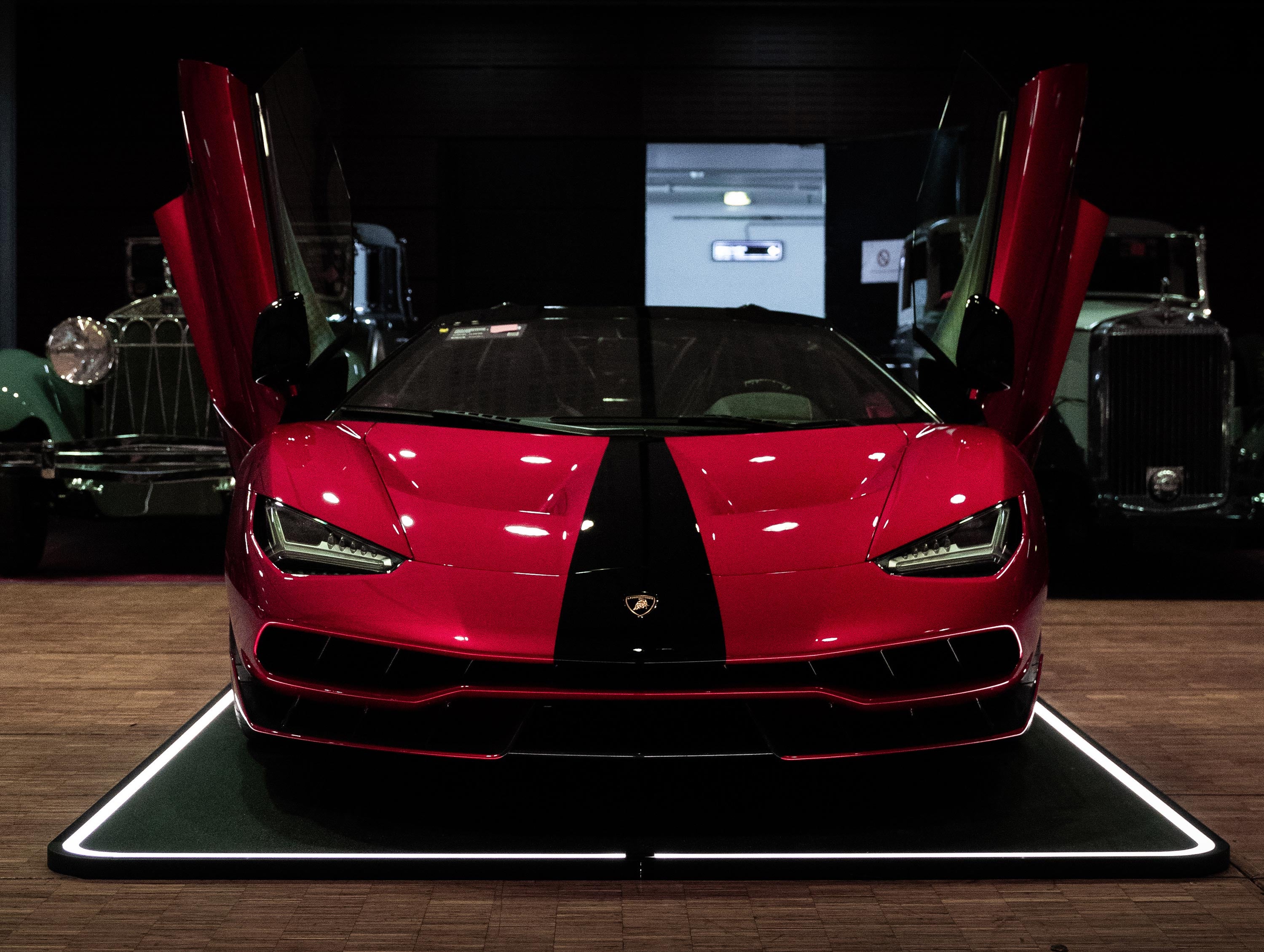 Roter Sportwagen mit offenstehenden Flügeltüren auf einem beleuchteten Pad, umgeben von klassischen Autos.