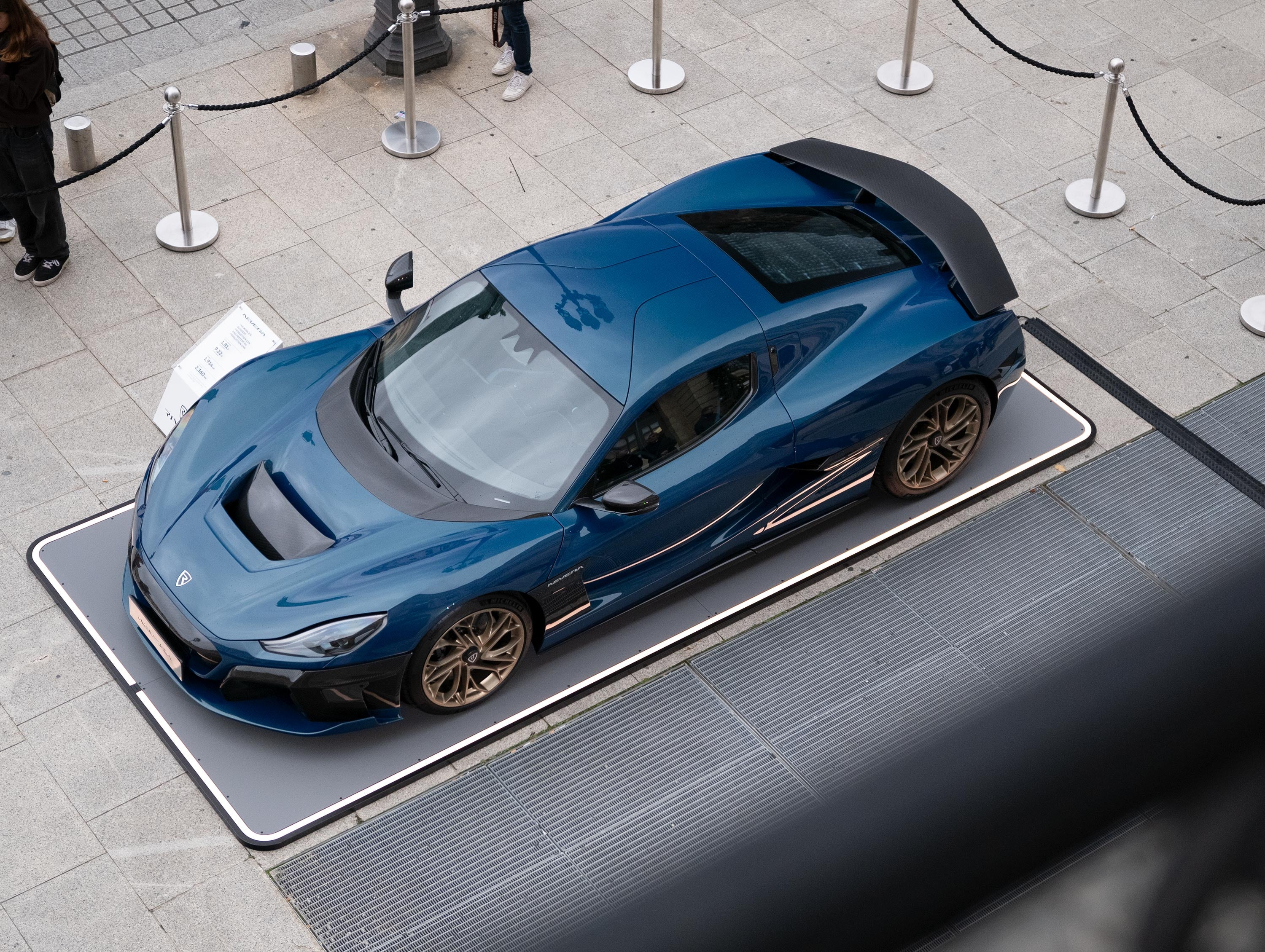 Blaues Auto auf rechteckigem Pad, umgeben von Absperrungen, auf einer gepflasterten Fläche ausgestellt.