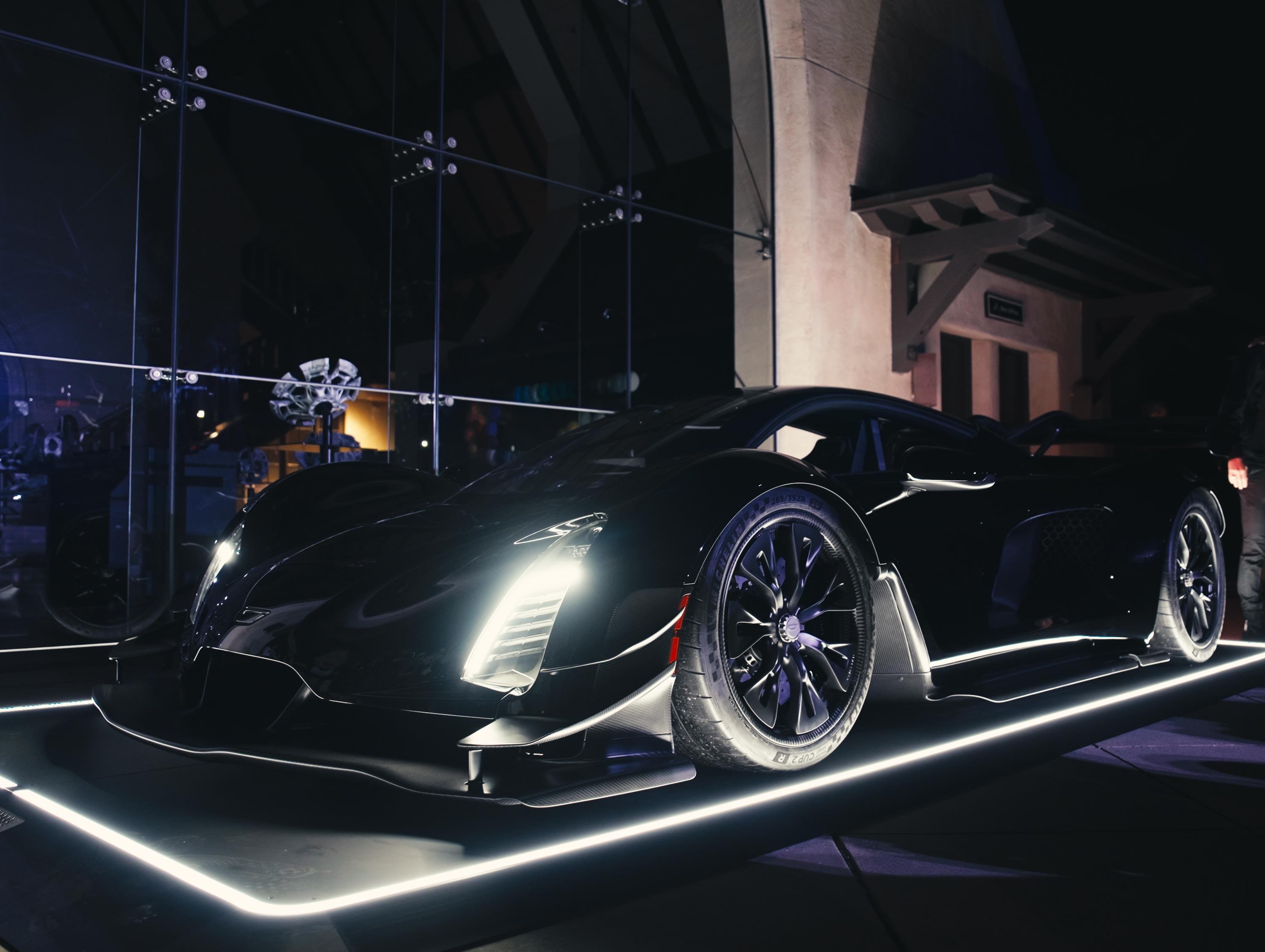 Ein schwarzes, sportliches Auto mit hellen Scheinwerfern steht nachts vor einem modernen Gebäude mit Glasfassade.