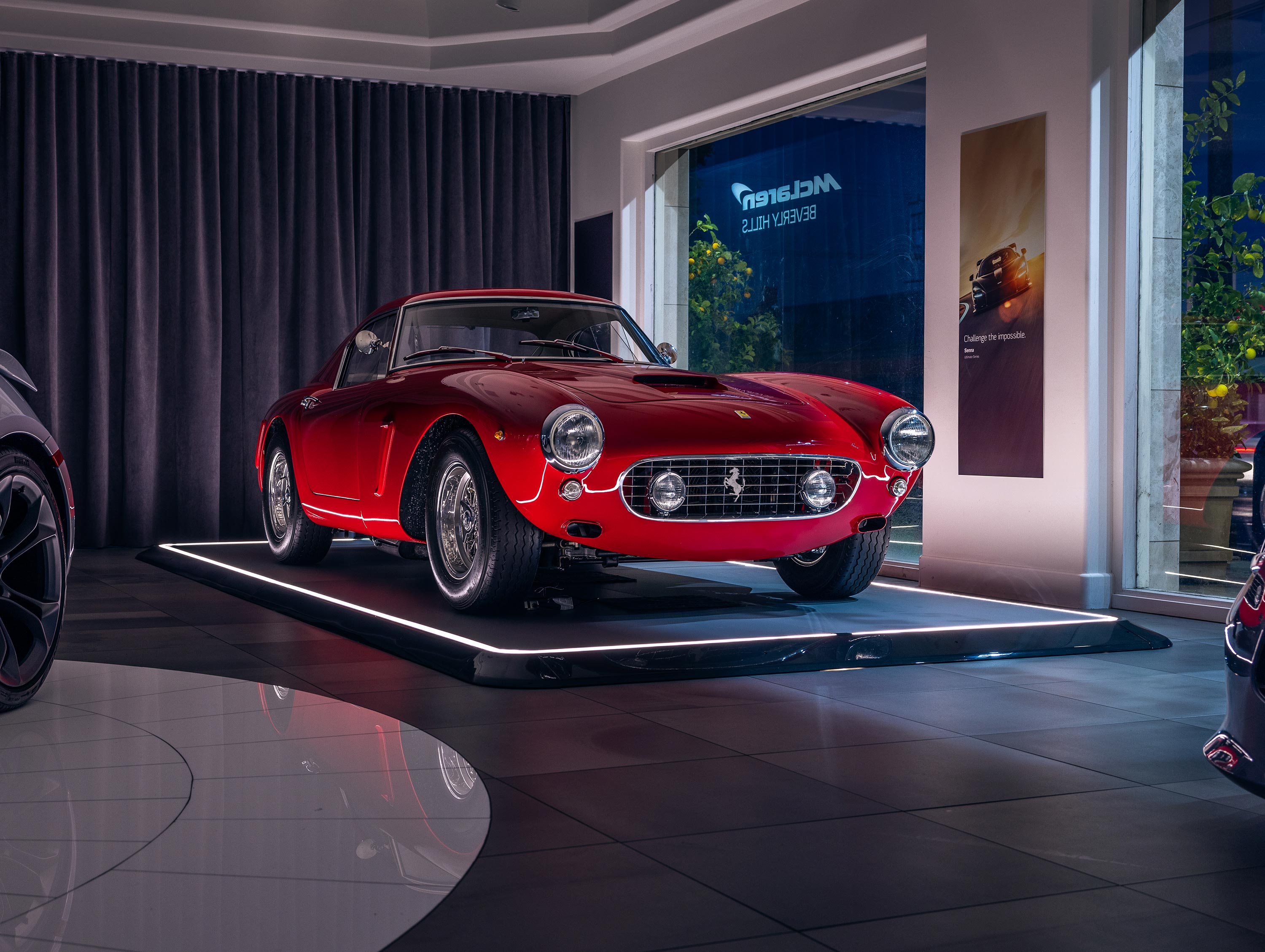 Roter Oldtimer-Sportwagen auf beleuchtetem Podium in einem eleganten Ausstellungsraum mit Vorhängen und großen Fenstern.