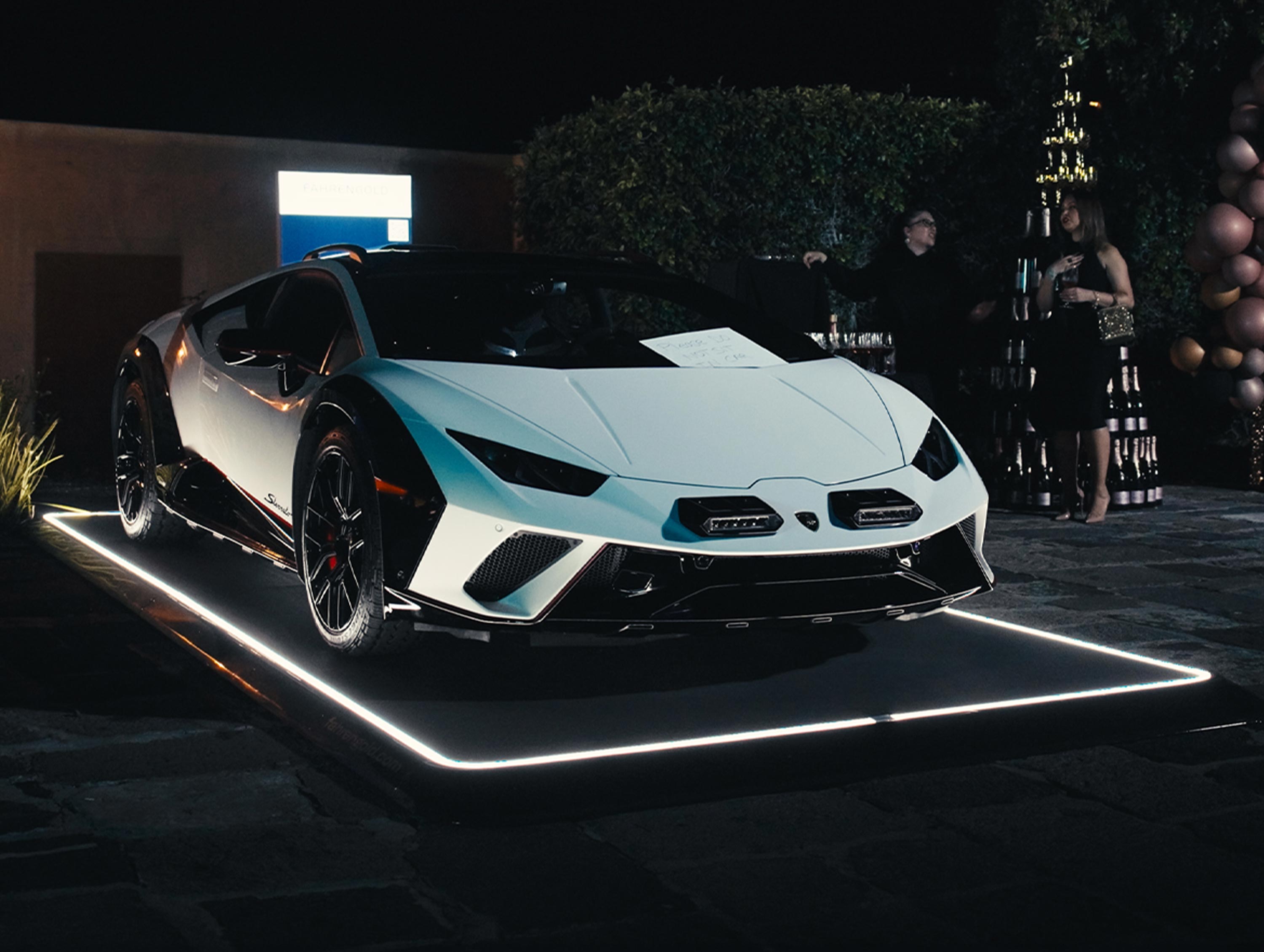 Ein weißer Sportwagen steht nachts auf einem beleuchteten Fahrzeugpad, umgeben von Menschen und Ballons.