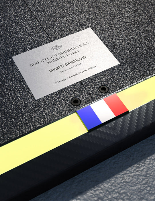 Metallplatte mit Gravur "Bugatti Automobiles S.A.S." auf rauer Oberfläche, darunter französische Flagge.