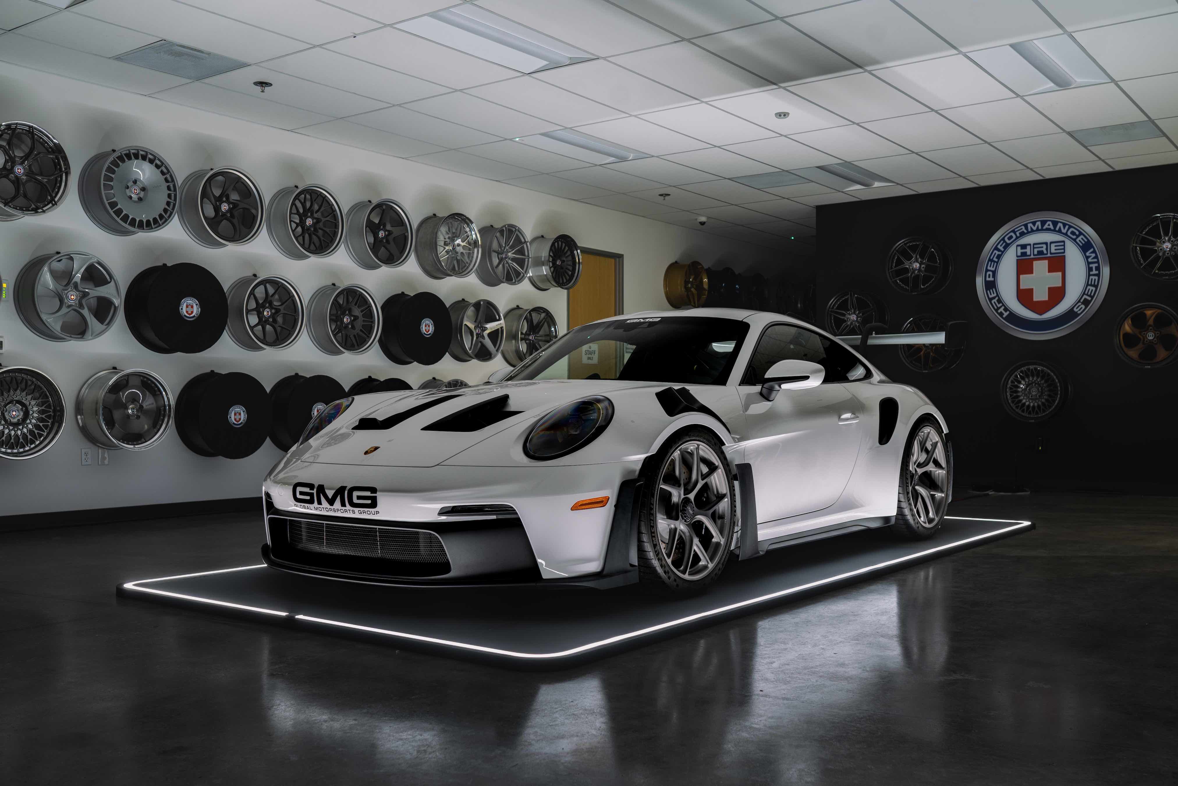 Weißes Auto auf beleuchtetem Pad in einem Raum mit vielen Autofelgen an der Wand.