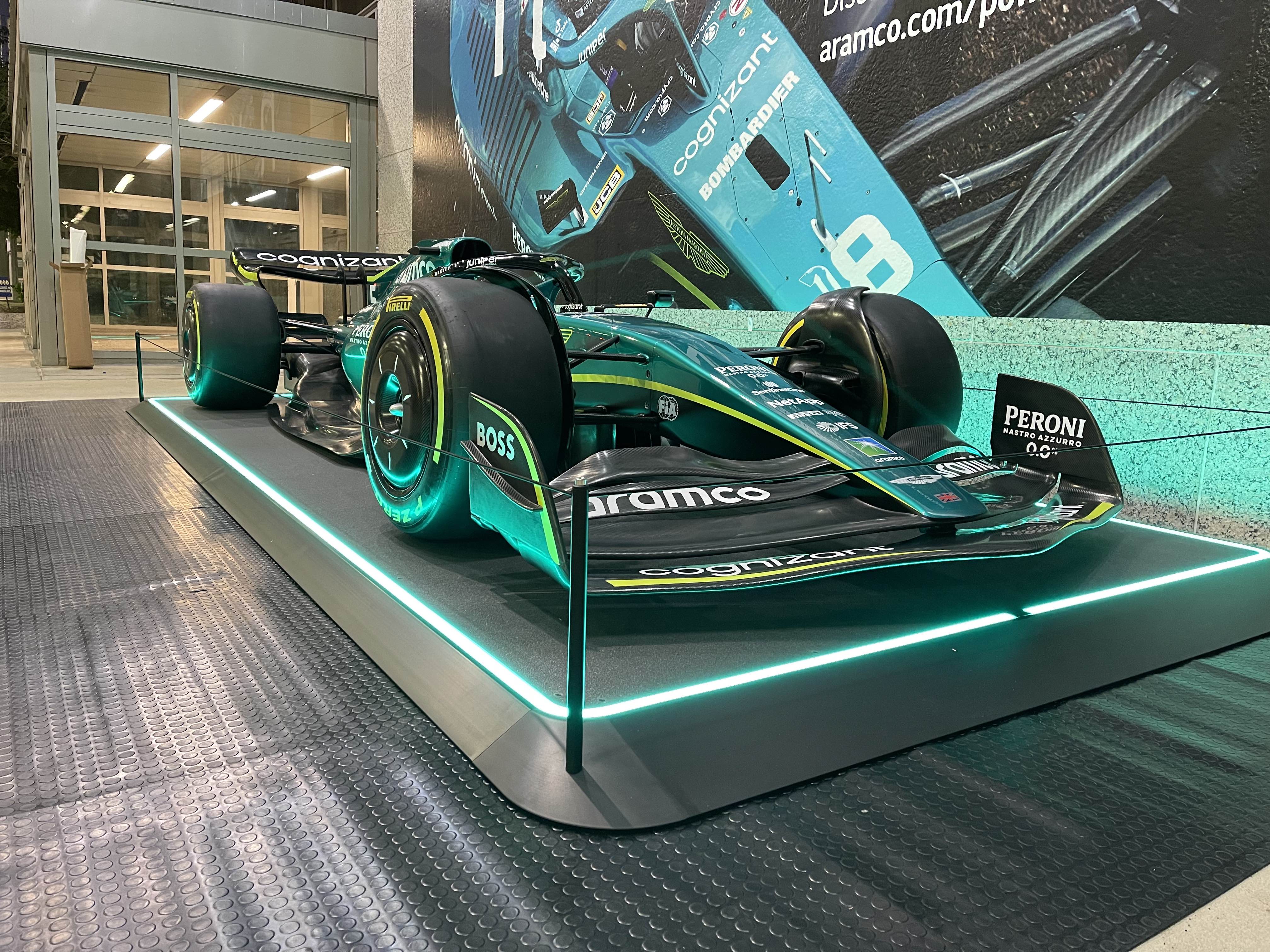 Rennwagen in auffälliger Beleuchtung auf einer erhöhten Plattform, mit Werbebildern im Hintergrund, nachts ausgestellt.