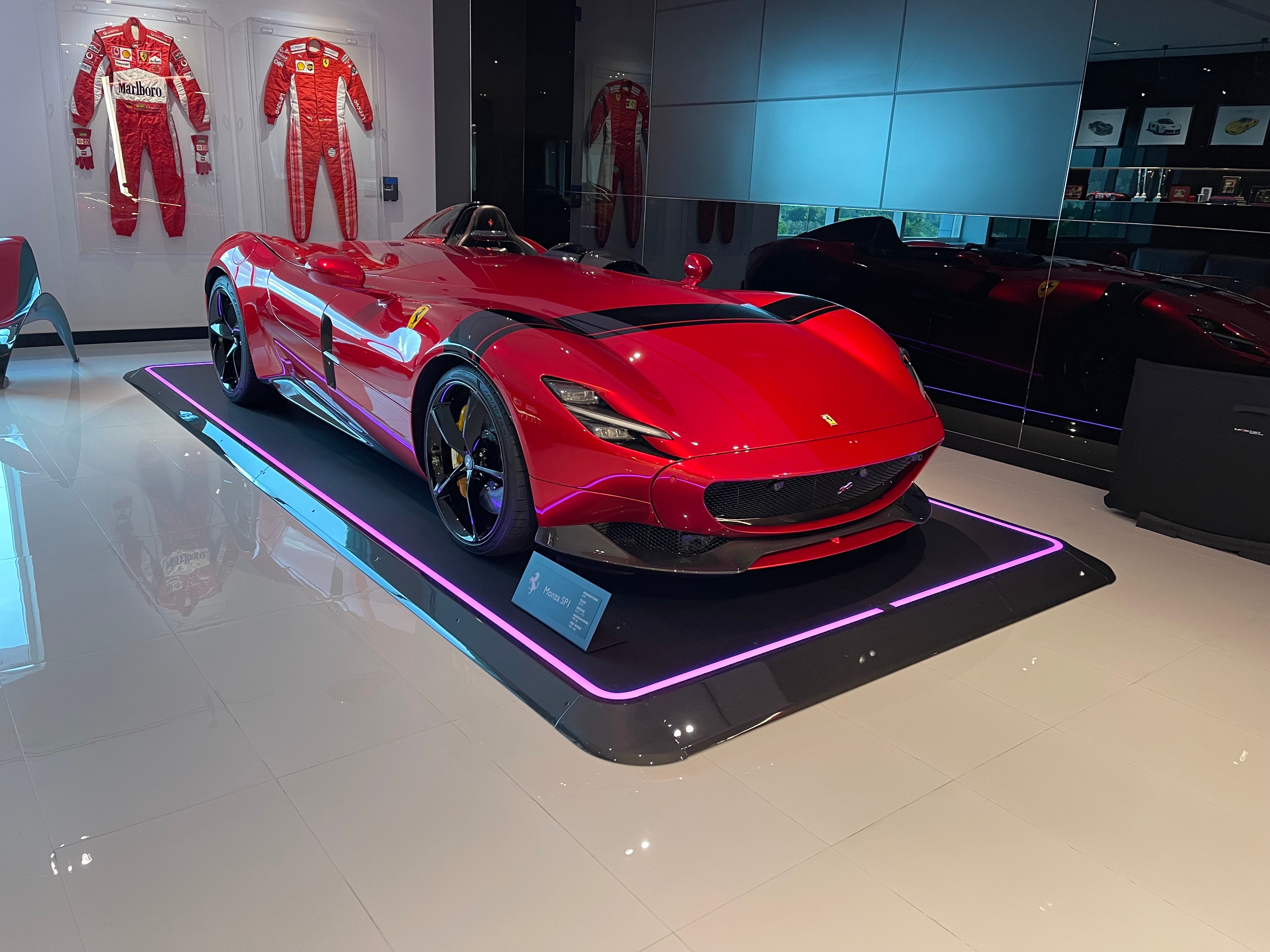 Ein rotes, sportliches Auto steht auf einem beleuchteten Pad in einem Ausstellungsraum mit Rennanzügen an den Wänden.