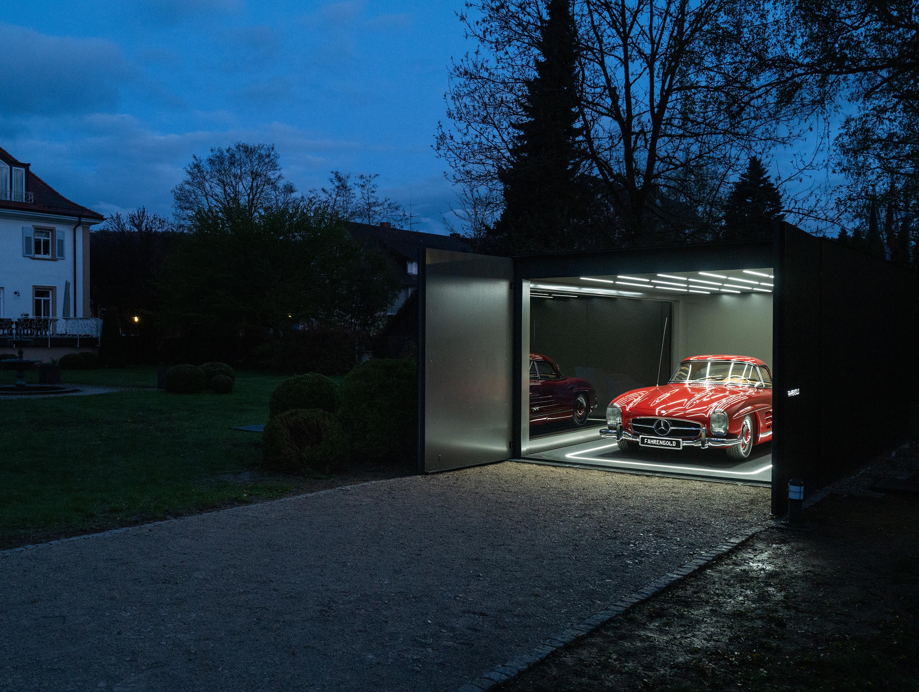 Zwei Oldtimer-Autos stehen in einer beleuchteten, modernen Garage bei Dämmerung, rechts ein klassischer roter Wagen.