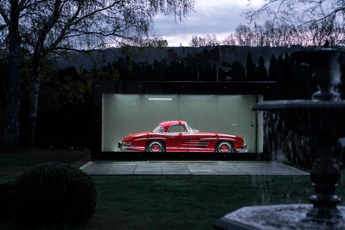Ein rotes Auto steht in einer modern beleuchteten Glasgarage, umgeben von Bäumen bei Dämmerung.