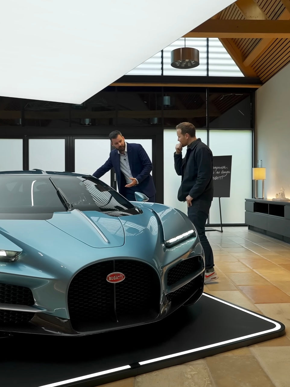 Zwei Männer stehen neben einem blauen Sportwagen auf einem rechteckigen Pad in einem modernen Raum mit Holzdecke.