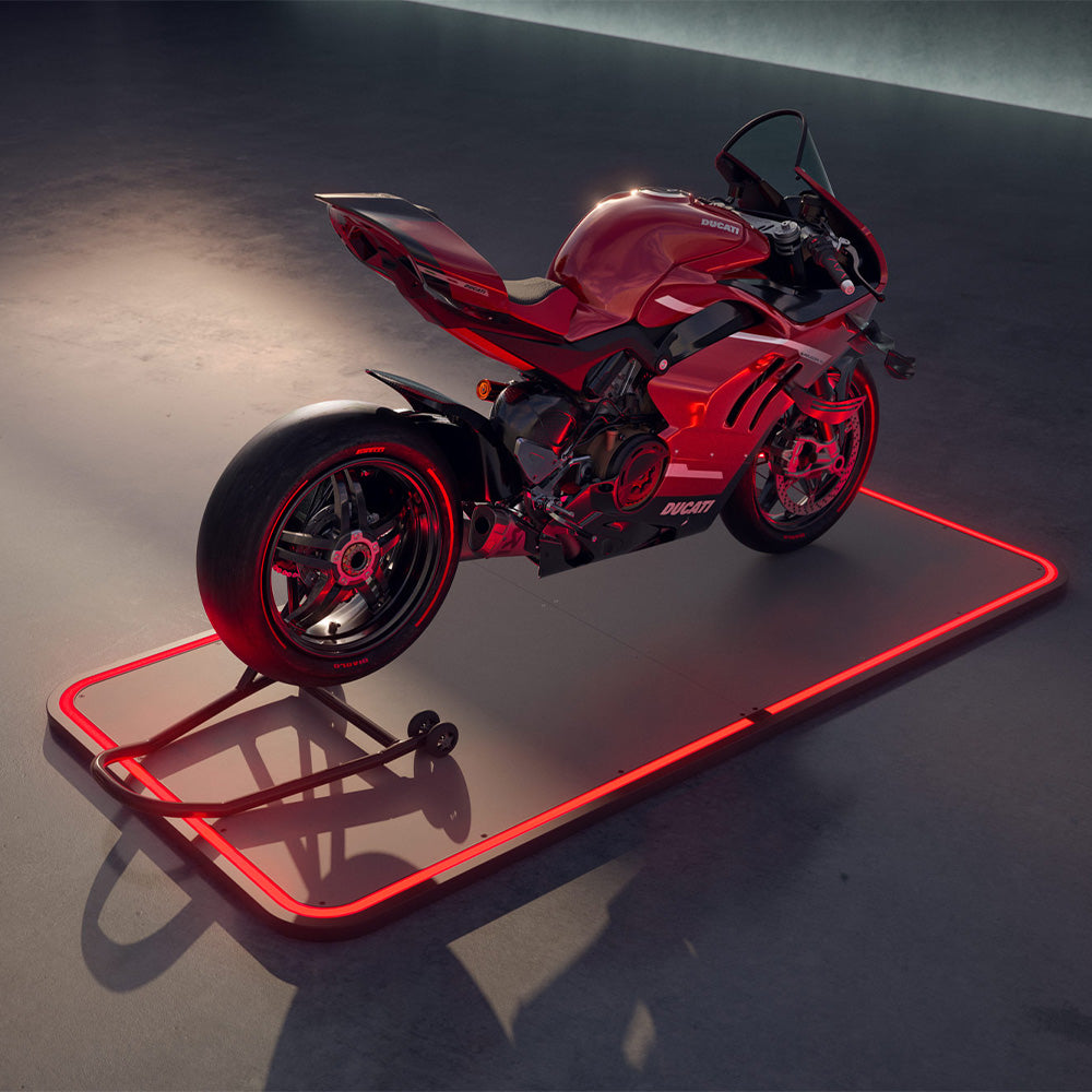 Rotes Motorrad steht auf beleuchtetem, rechteckigem Pad auf grauem Boden in abgedunkelter Umgebung.