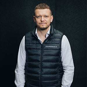 Eine Person mit gestreifter Weste und weißem Hemd steht vor einem dunklen Hintergrund.