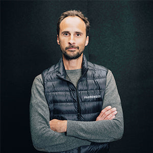 Mann mit Bart, grauem Pullover und schwarzer, gesteppten Weste vor schwarzem Hintergrund mit verschränkten Armen.