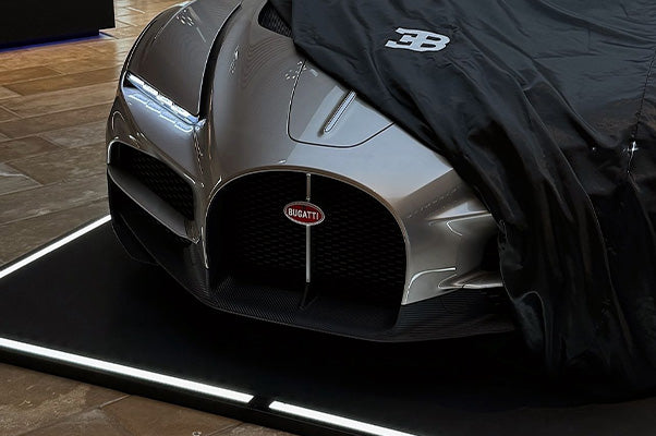 Ein teilweise abgedecktes graues Auto mit einem "Bugatti"-Emblem steht auf einem beleuchteten schwarzen Pad.